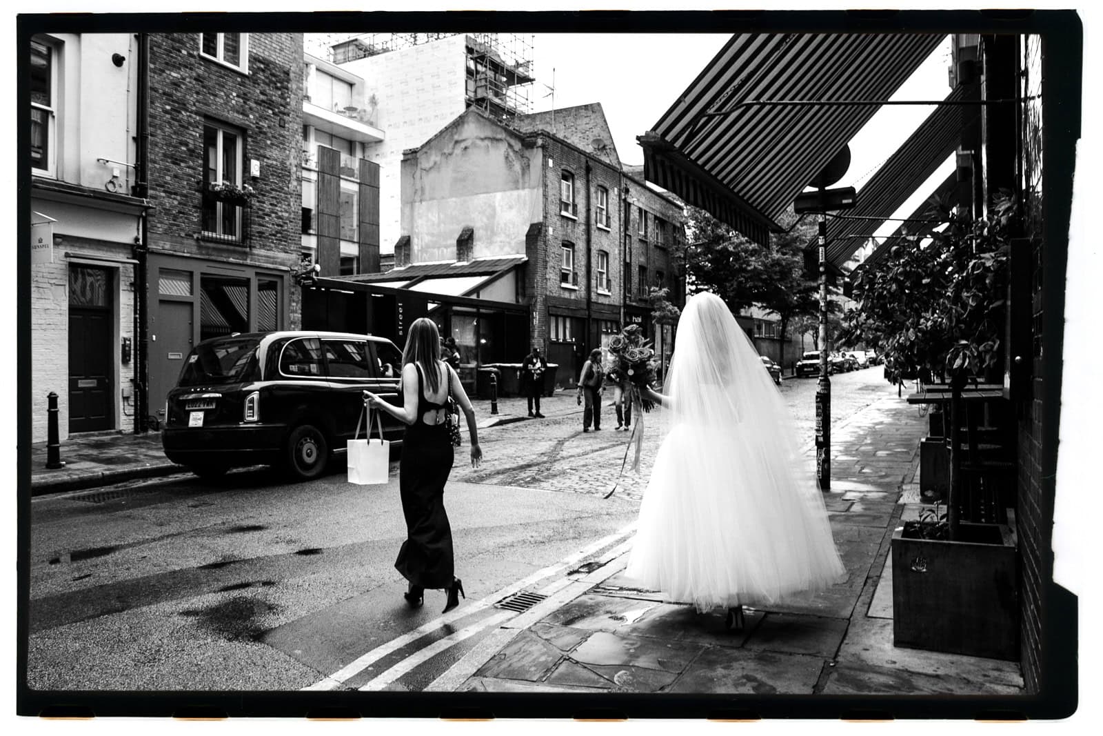 A bride and a guest walk through a London street, with the full black film rebate visible