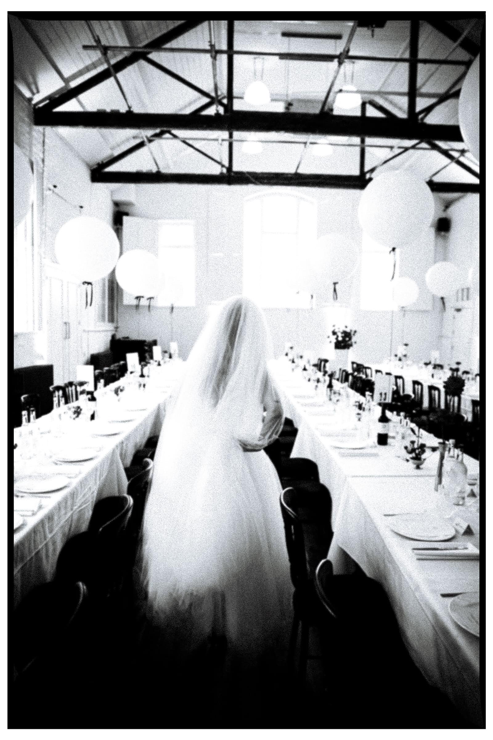 A bride walks between long tables in a bright reception room, photographed in black and white with a dark border