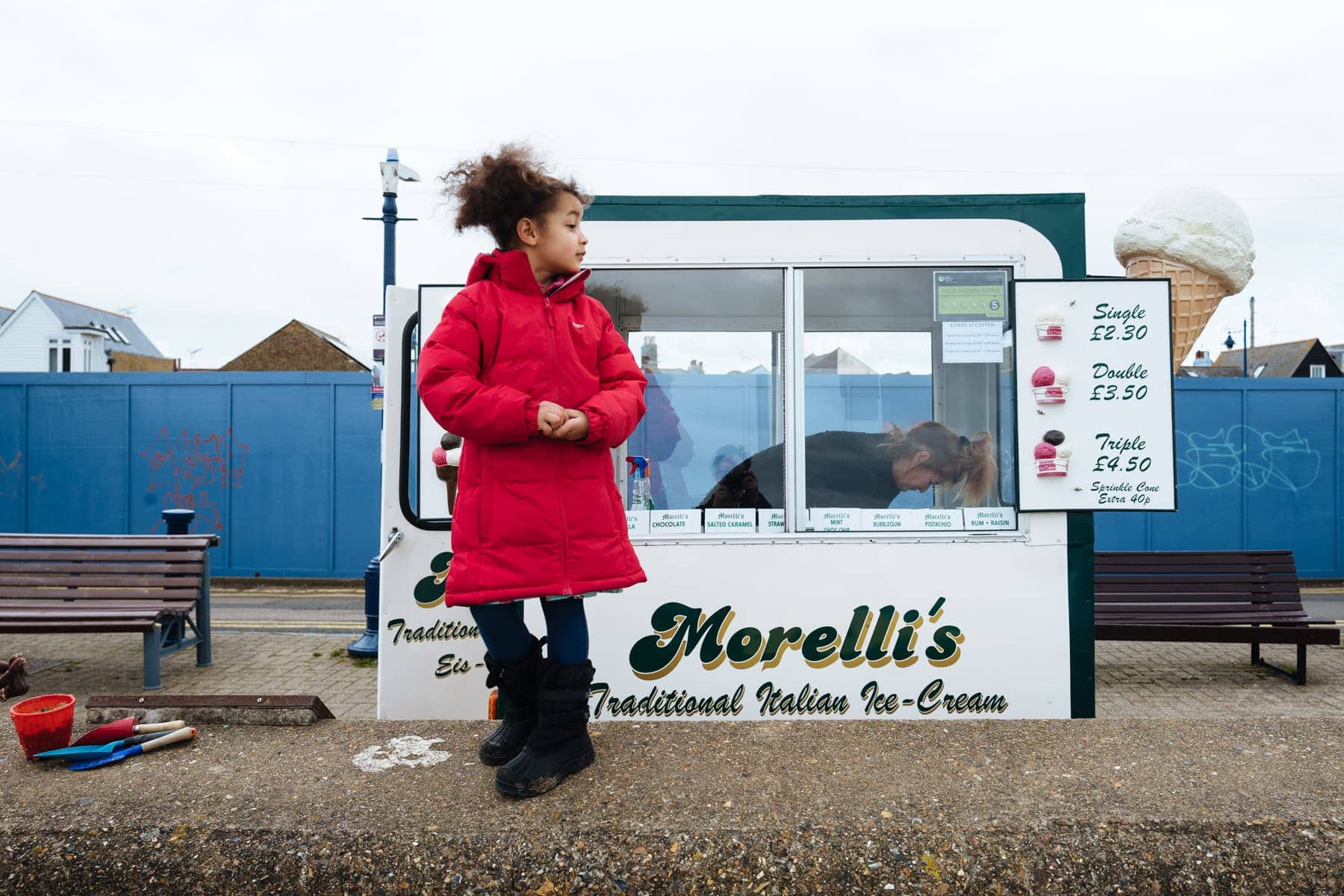 A child in a red coat stands beside a seaside ice cream hut