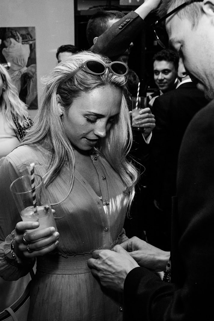 A guest fastens another guest’s dress in a crowded room
