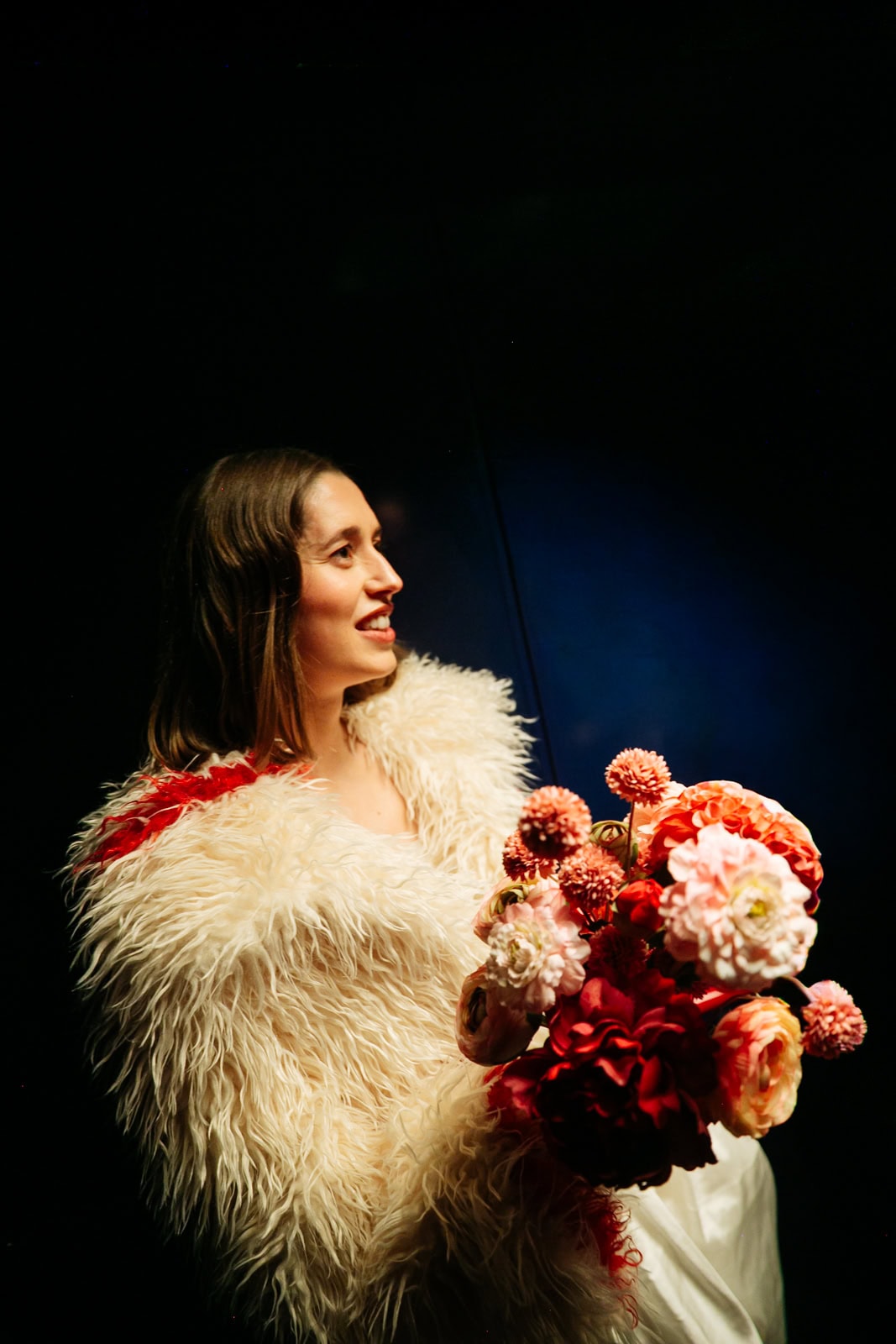 A bride in a white fur coat holds flowers against a dark backdrop