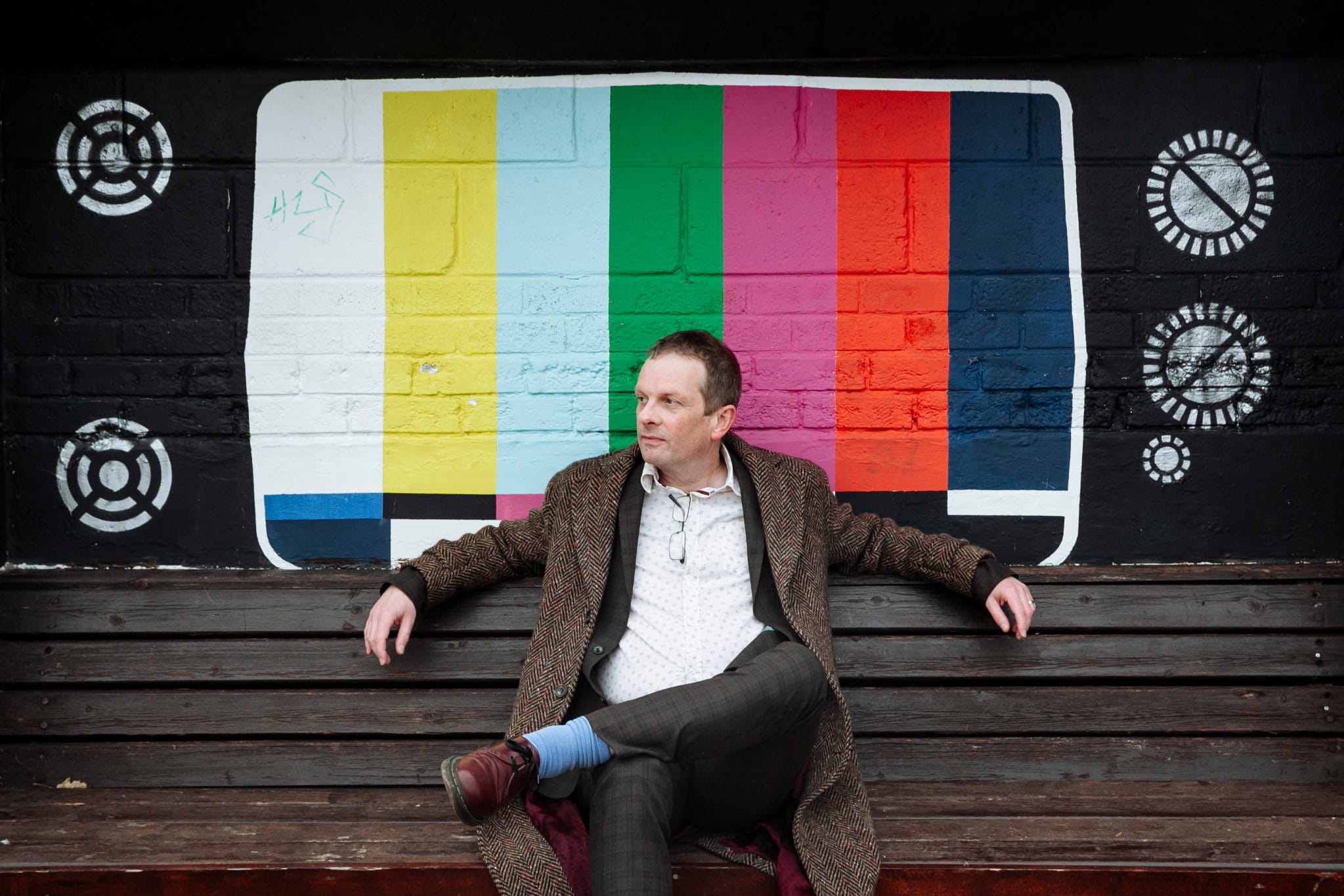 Author John Higgs seated in front of a television test-card mural.