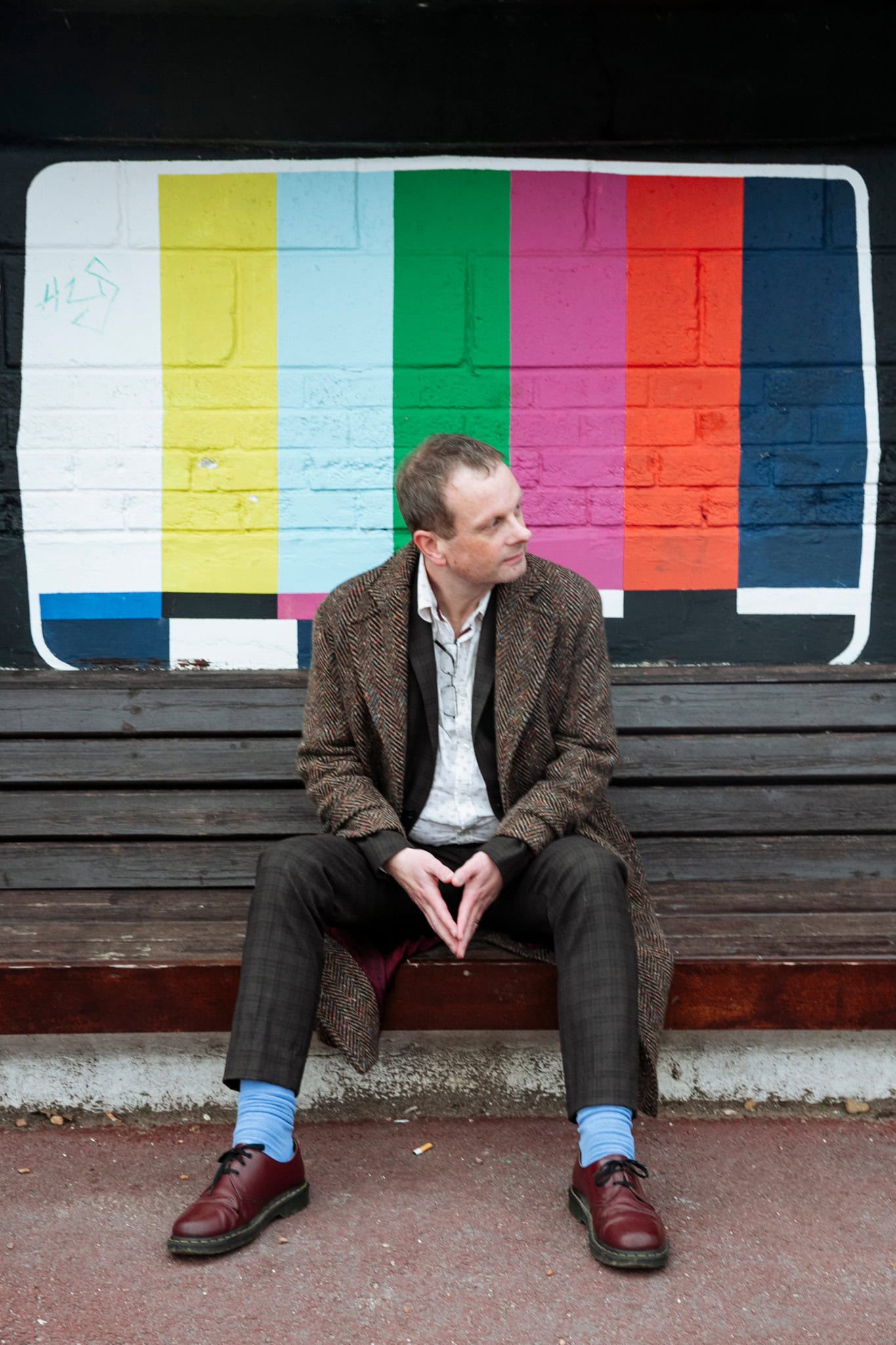 Author John Higgs seated on a bench beneath a colourful television test-card mural, looking to one side.