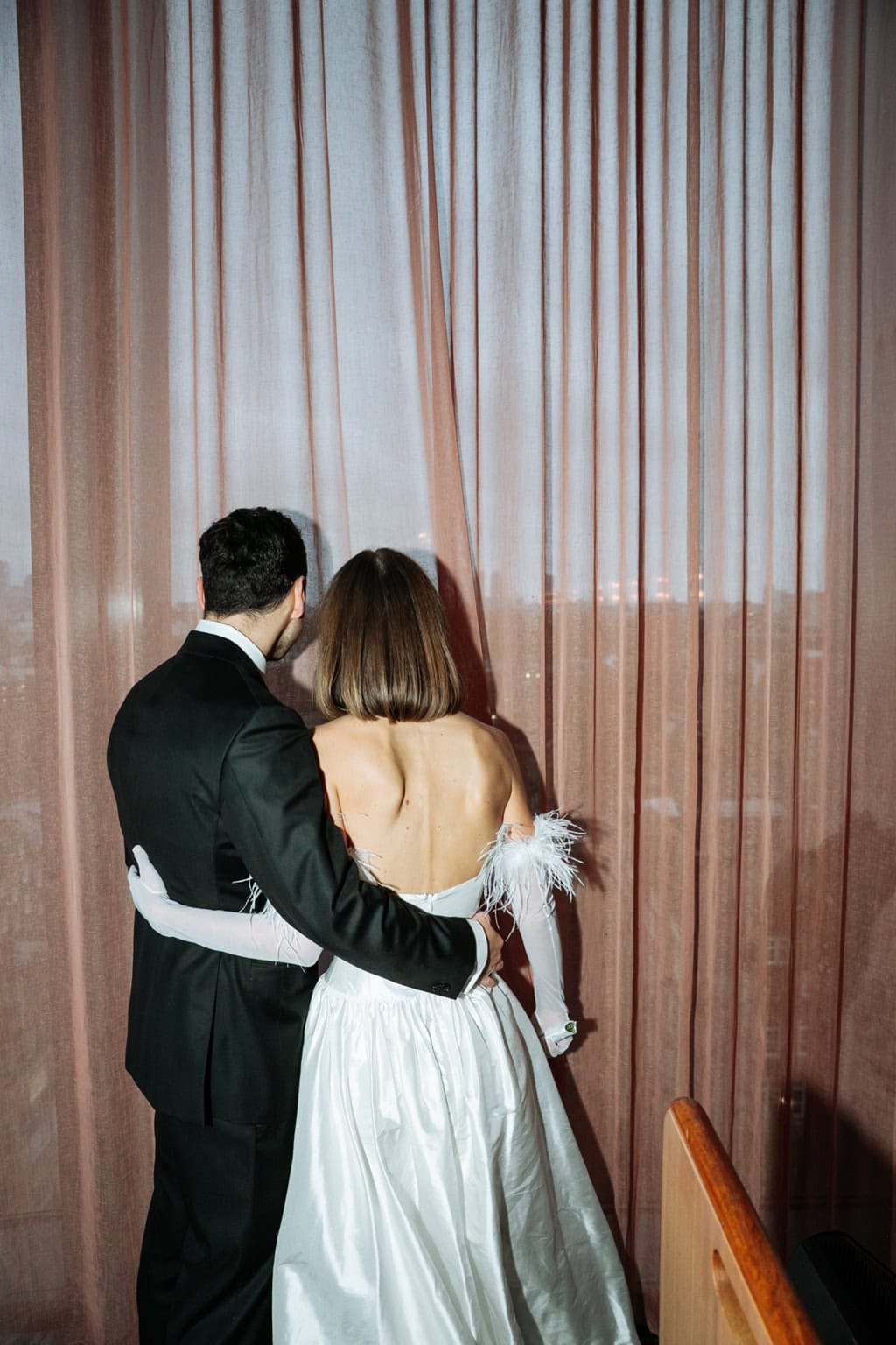 A couple stands together facing pale curtains