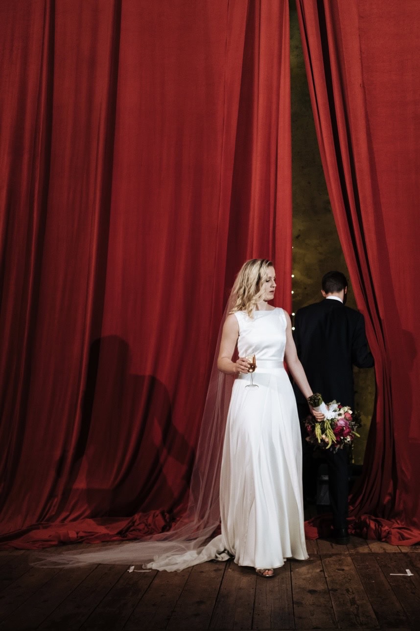 A bride and groom stand before a huge red curtain