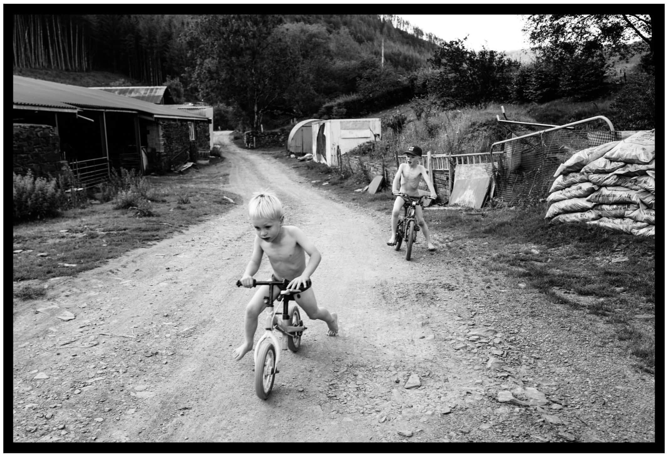 A small child pedals a bicycle down a rough lane while another follows